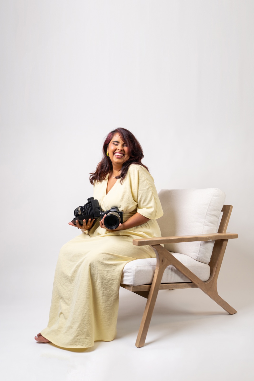 Tanya Chopra laughing, seated in a chair, holding a vintage film camera and a Sony mirrorless.